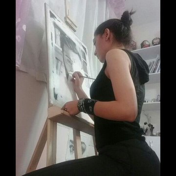 A woman in a black dress with a wristband paints a portrait of a face on a canvas at an easel in a sunlit studio.