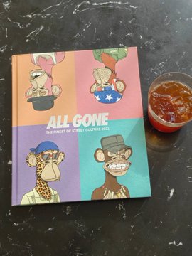 The image features a book titled "All Gone" with cartoon illustrations of various characters, including a monkey wearing sunglasses. The book appears to be a limited edition item, as indicated by the text "NFT之夏那年". A cup is placed next to the book on a table, suggesting that someone might have been enjoying a drink while reading or admiring the artwork in the book.