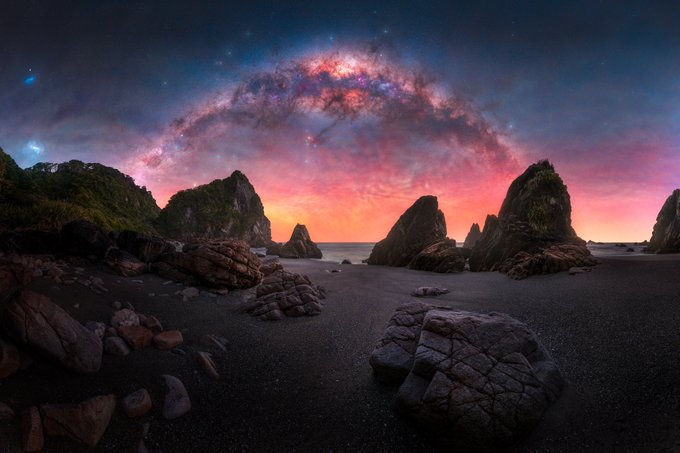 A stunning astrophotography landscape featuring a vibrant, arching Milky Way galaxy over a rocky coastline at sunset, with dramatic pink and orange hues in the sky.