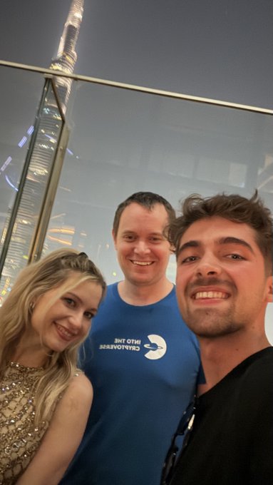 A selfie of three smiling people posing together at night with the Burj Khalifa visible in the background. The man in the middle wears a blue "Into The Cryptoverse" t-shirt, suggesting they are at a crypto or Bitcoin-related event, aligning with the post's mention of Benjamin Cowen's upcoming Miami conference about teaching kids and teens about Bitcoin and compounding interest.