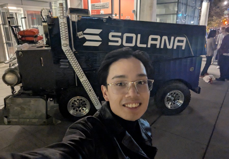 A young man wearing glasses takes a selfie in front of a blue street sweeper truck with a large 'SOLANA' logo on the side.