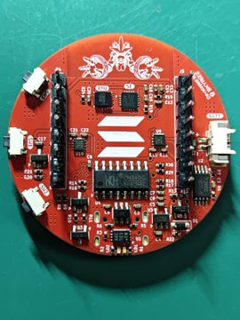A close-up photograph of a red circular SparkFun development board (likely a MicroMod or Qwiic board) with visible microchips, connectors, and white logos printed on the PCB.