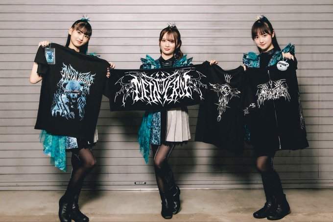 Three young women in dark, gothic-style outfits with teal accents pose together while holding up four black band t-shirts featuring intricate metal-style logos and designs.