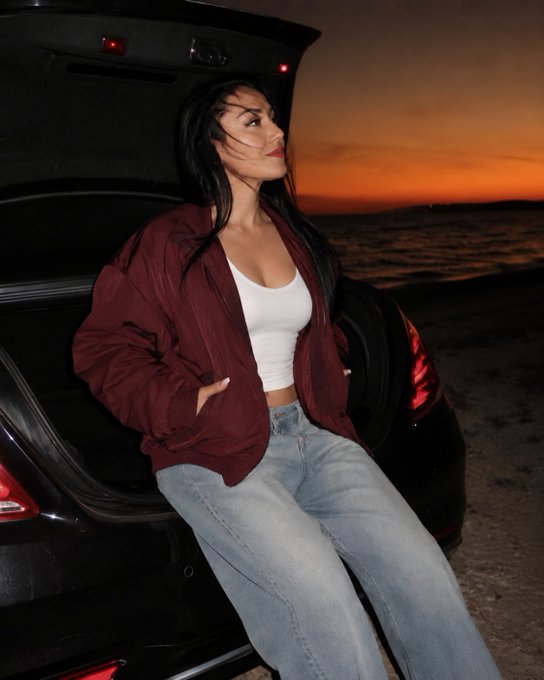 A woman with long dark hair wearing a maroon bomber jacket, white crop top, and light blue jeans leans against the open trunk of a black car at sunset near the ocean.
