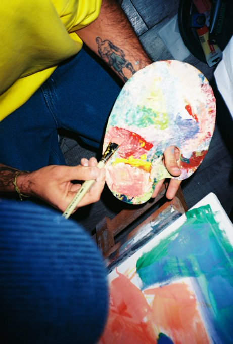 An overhead view of an artist's hands holding a paint-stained palette and brush while working on a colorful abstract painting, with tattoos visible on their forearm.
