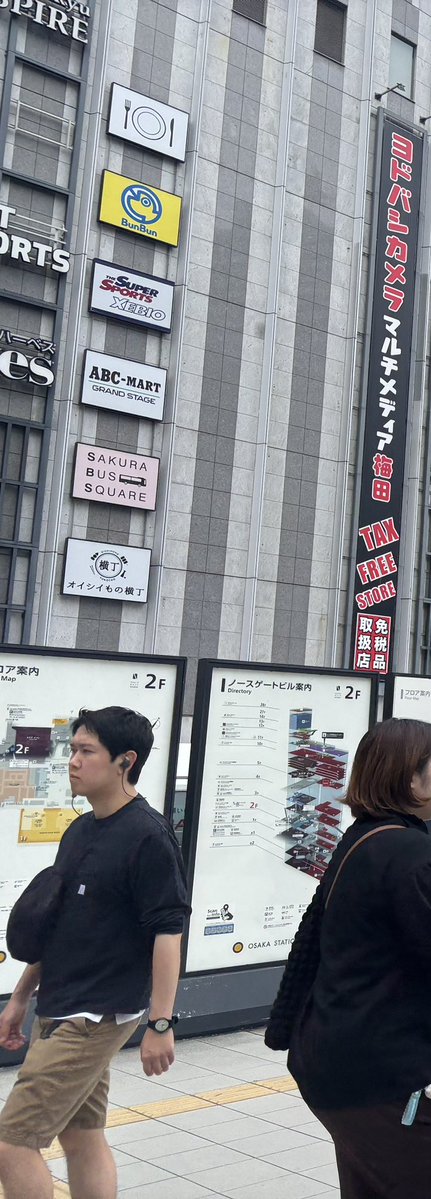 A street scene outside a building in Osaka with signs for 'TAX FREE STORE', 'BunBun', 'Super Sports XEBIO', 'ARC-MART GRAND STAGE', and 'SAKURA BUS SQUARE', where a man in a black shirt and a woman are walking past a directory map.