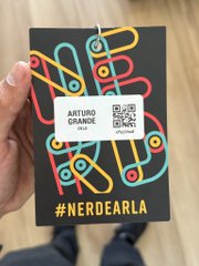 A hand holds up a black event badge with colorful linked graphics, displaying the name "ARTURO GRANDE" and the hashtag "#NERDEARLA."