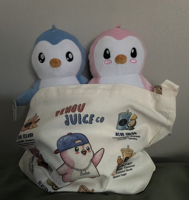 Two penguin plush toys (one blue, one pink) sit behind a "PENGU JUICE CO" canvas tote bag featuring cartoon graphics.