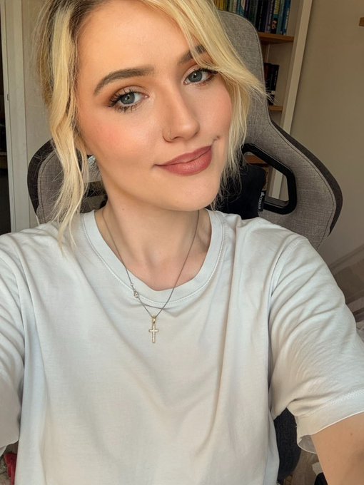 A blonde woman with a soft smile wears a white t-shirt and layered necklaces, including a gold cross pendant, sitting in a gray gaming chair with a bookshelf visible behind her—presenting a cozy, ready-for-work vibe perfect for a "gm" morning check-in.