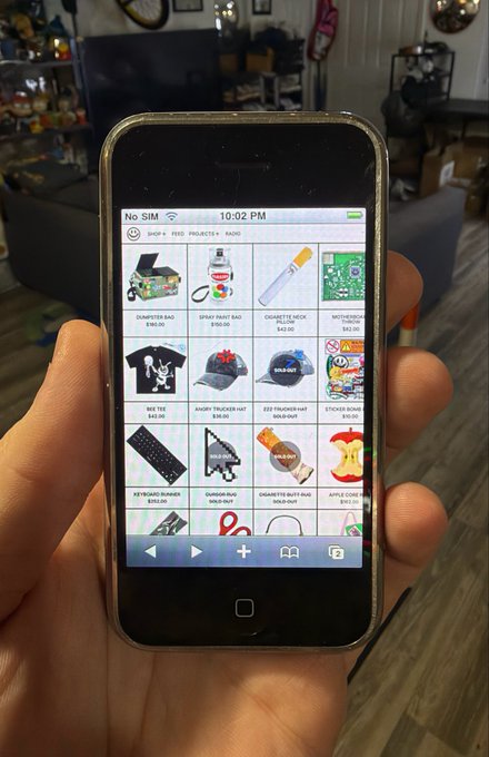 A hand holds an old iPhone displaying an online shop app with various smoking accessories and branded merchandise for sale, including items like a "Smart Bait Bag," "Djarum Black Cigarette Holder Pillow," and an "Apple Core Rolling Tray." The phone's status bar shows "No SIM" and the time "10:02 PM," with navigation tabs for "Shop," "Feed," "Projects," and "Raid" visible at the top.