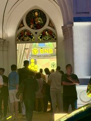 A group of people stands in line inside an ornate venue with Gothic arched stained-glass windows, looking toward a brightly lit yellow screen in the distance.
