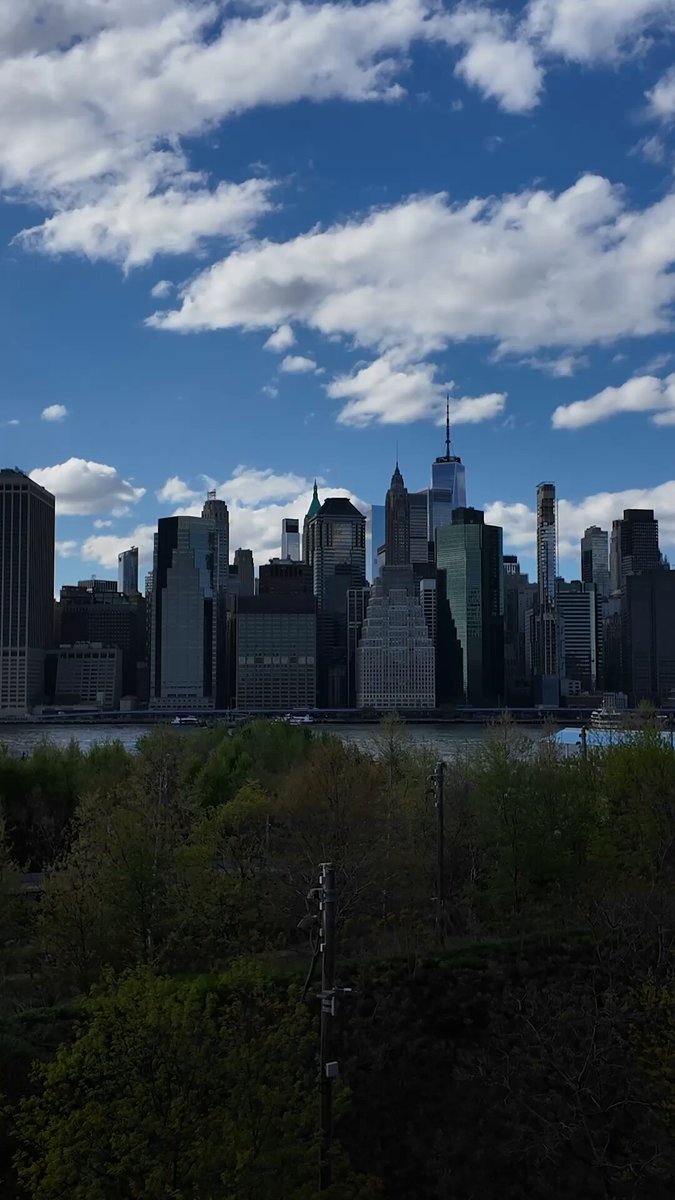 The image shows a city skyline with tall buildings, including the New York City skyline, which includes the Empire State Building. The sky above the city has white clouds scattered throughout. In the foreground of the photo, there are trees that add greenery to the scene. The caption reads "Yale, NYU, Fordham, Columbia, Cornell Tech." and "9 days. 5 campuses. 3 workshops alongside @1inch".