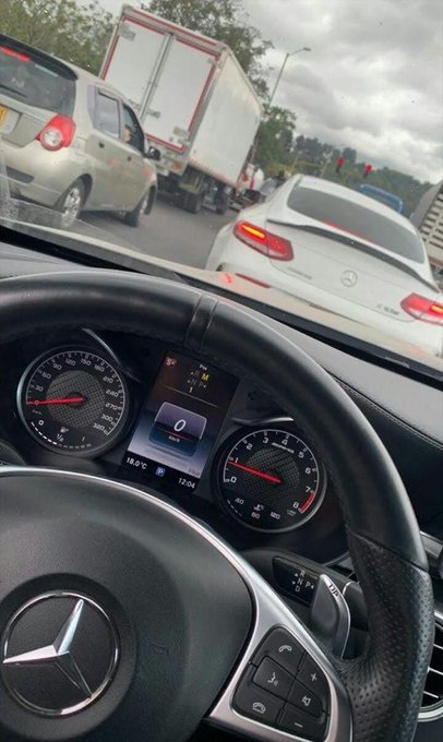 The image shows a view from behind the steering wheel of a Mercedes-Benz, with the dashboard displaying 0 mph, 18.5°C, and 12:04, while stopped in traffic alongside a white Mercedes C-Class sedan. The post's "GM" greeting (meaning "good morning") pairs with the morning commute scene captured here, suggesting the driver is starting their day "First light first win" during a traffic-filled "Thirsty Thursday."