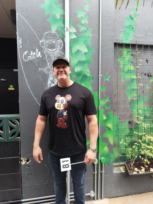 A man wearing a "Killa" bear t-shirt and cap poses in front of a gray wall with green ivy and a surfboard mural reading "Catch," holding a sign numbered "81," fitting the post about showing off his Killa Bear NFT drip.