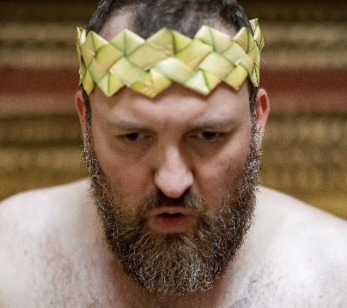 A close-up image of a bearded man wearing a woven green leaf crown, looking downward.