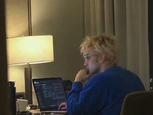 A blonde-haired person wearing glasses and a blue sweatshirt sits thoughtfully at a desk in front of an open laptop displaying what appears to be trading data, illuminated by a table lamp.