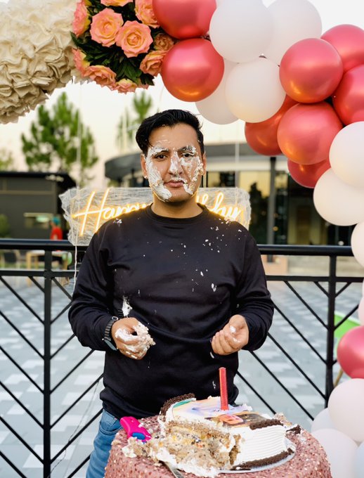 A man stands with a smashed birthday cake in front of him, wearing a black shirt covered in frosting, with a "Happy Birthday" neon decoration and balloon arch behind him. The accompanying post celebrates his birthday, expressing gratitude for life, family, and community—matching the festive, joyfully chaotic scene of a cake being playfully destroyed.