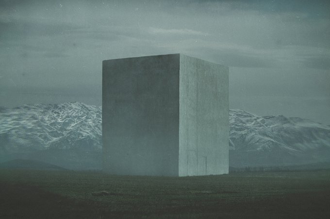 A large concrete cube standing in a grassy field against a backdrop of snow-capped mountains under a cloudy sky.