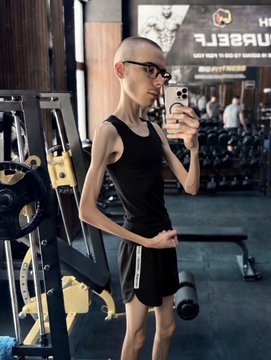 A young person with a shaved head and glasses takes a mirror selfie in a gym, wearing a black tank top and shorts while holding a phone.