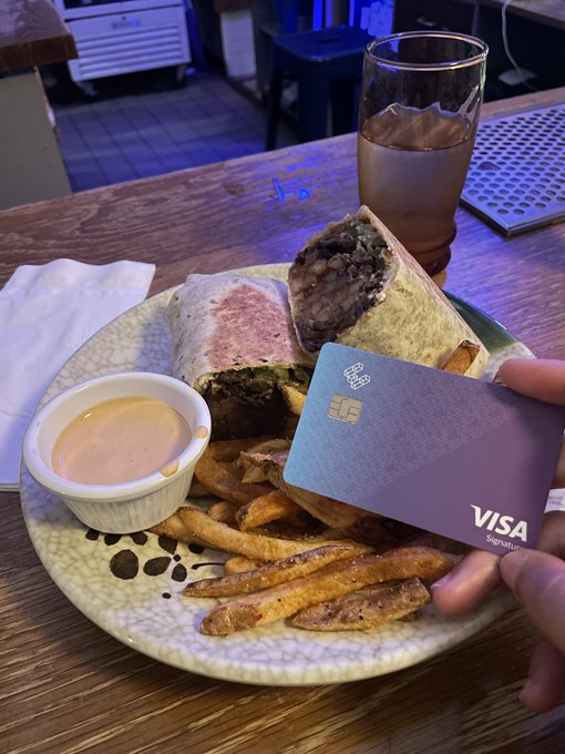 A Visa Signature credit card is held over a plate featuring a burrito sliced open to reveal bulgogi-style meat, crispy fries, and a dipping sauce, with an iced drink in the background. The image ties to the caption by illustrating everyday real-world purchases, suggesting that even items like a Korean California Burrito could be bought with stablecoins in place of traditional payment methods.