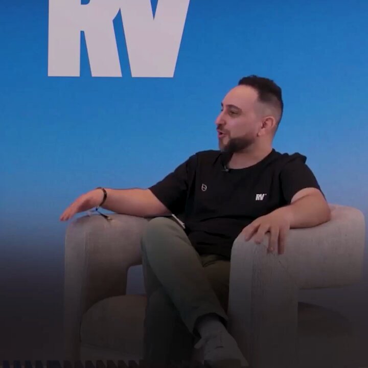 The image features a man sitting comfortably in an armchair with his legs crossed while wearing a black shirt and green pants. The background of the image shows a blue sky, which contrasts nicely with the white text that reads "RV". There is also some text on the right side of the image, but it appears to be unrelated to the main subject - the man in the chair.