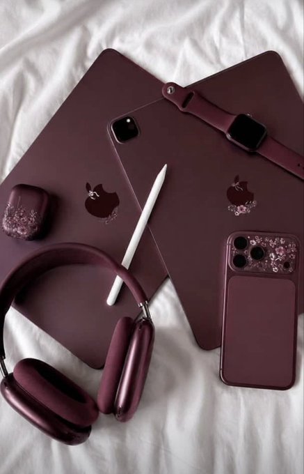 A flat-lay arrangement of matching burgundy/maroon Apple products including an iPad, iPhone, Apple Watch, AirPods case, AirPods Max, and an Apple Pencil on white sheets.