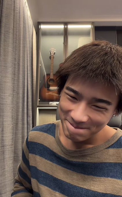 A smiling young man with short brown hair wearing a striped sweater, photographed indoors with an acoustic guitar visible in the background.