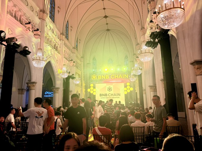 A crowded event in a grand hall with chandeliers and arched ceilings, featuring a stage with a large illuminated 'BNB CHAIN SUPER REGULAR SINGAPORE' sign.