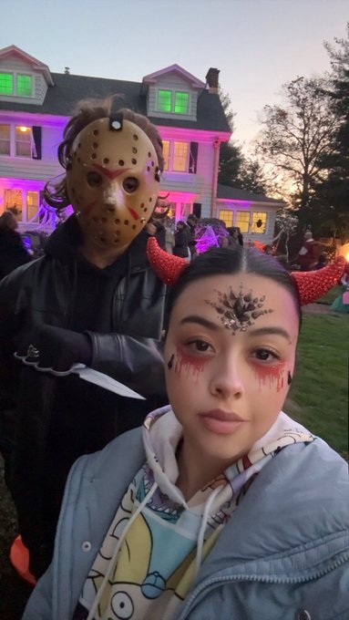 A selfie of a couple in Halloween costumes in front of a house decorated with purple and green lights; the man wears a Jason Voorhees hockey mask holding a bloody knife, and the woman has red horn headbands and bloody eye makeup.