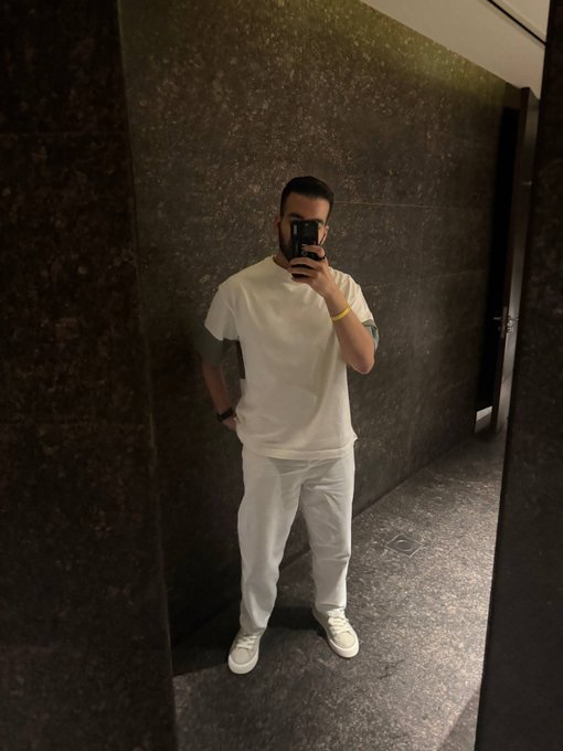A mirror selfie of a man wearing an all-white outfit consisting of a white t-shirt with gray sleeve accents, white pants, and white sneakers, standing in a dark marble-tiled bathroom or hallway while holding a phone.