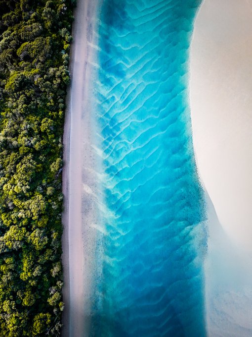 An aerial view shows a pristine white sand beach bordered by lush green tropical forest on one side and vibrant turquoise ocean water on the other.