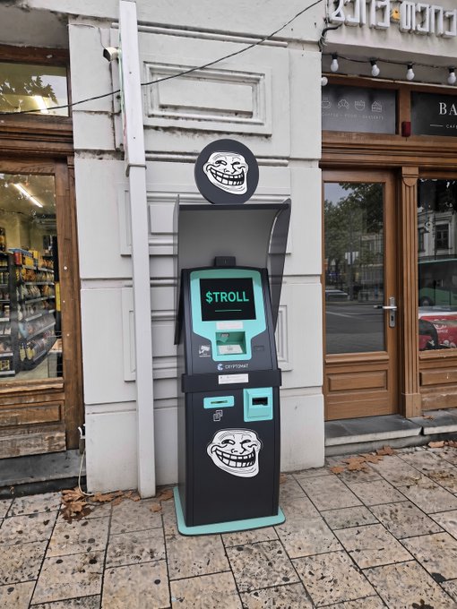 A '$TROLL' cryptocurrency ATM stands on a sidewalk outside a storefront, featuring the classic Trollface meme on both its top sign and lower body.