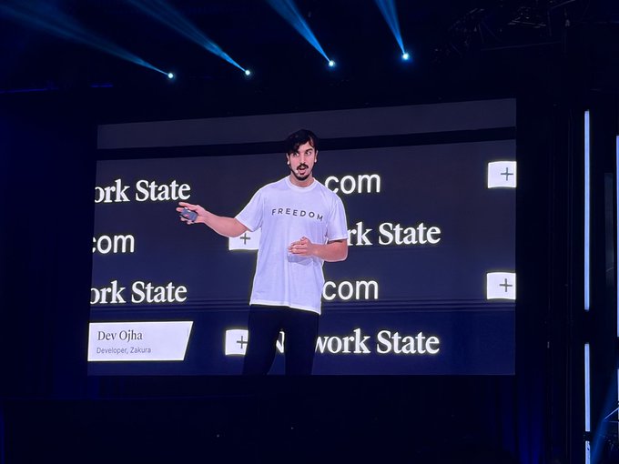 A man identified as 'Dev Ojha, Developer, Zakura' stands on stage giving a presentation, wearing a white t-shirt that reads 'FREEDOM' against a screen displaying 'New York State' branding.
