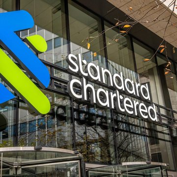 The image shows the exterior of a Standard Chartered bank building, featuring the company's logo with its signature blue and green cross-like emblem alongside the white "standard chartered" text. This relates to the post as Standard Chartered is the financial institution issuing the $10 price target for Arbitrum (ARB), highlighting the bank's involvement in crypto-related financial projections.