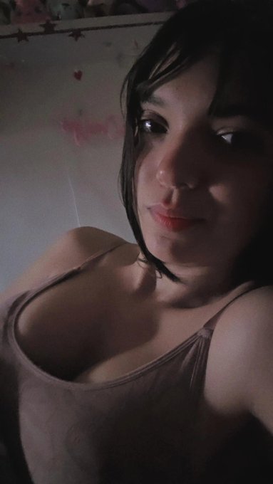 A dimly lit selfie shows a woman with dark hair and red lipstick wearing a beige tank top against a wall decorated with pink hearts and stars.