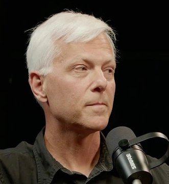 A close-up portrait shows a man with white hair wearing a black shirt, speaking into a black Shure microphone against a dark background.