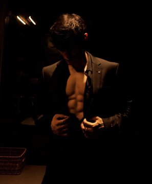 A man in a dimly lit setting wears a partially unbuttoned dark suit jacket, revealing his bare, muscular torso as he looks downward.