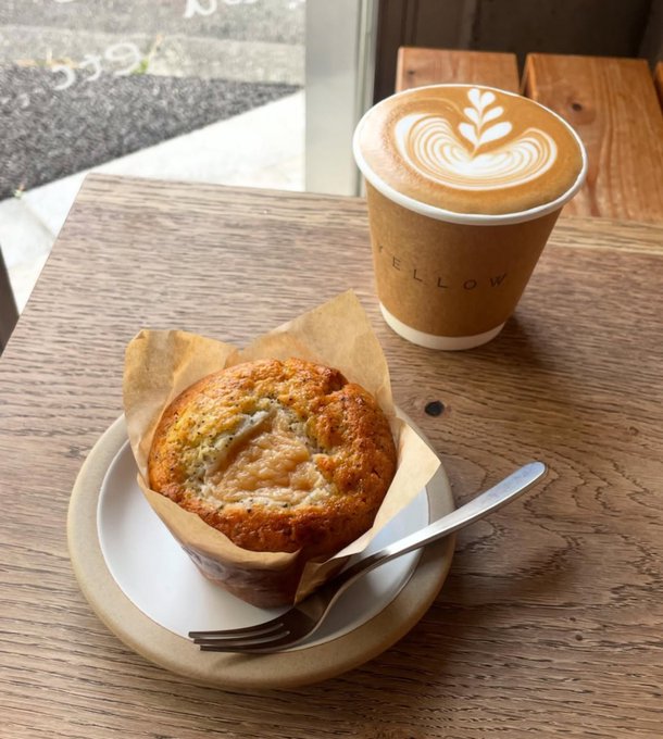 A coffee and muffin served on a wooden table at a cafe called 'YELLOW', featuring a latte with a leaf-shaped foam art design in a brown paper cup alongside a golden-brown muffin on parchment paper with a fork on a beige plate.