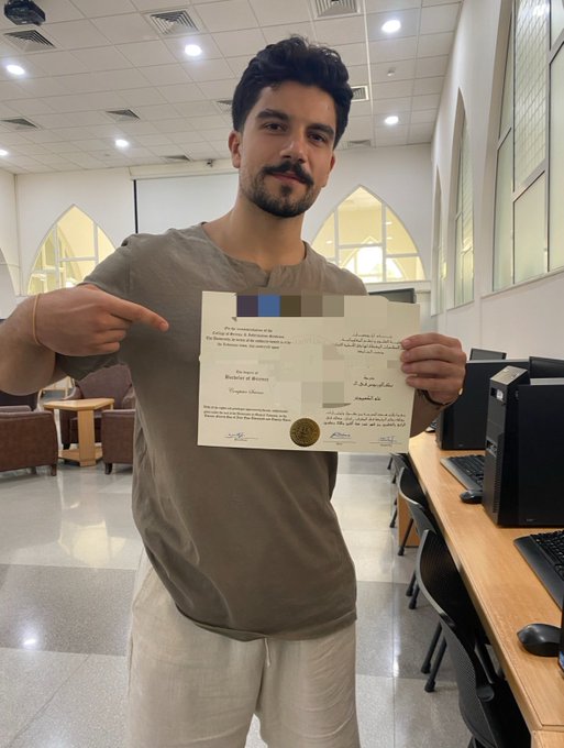 A young man stands in a computer lab, smiling and pointing at a certificate he is holding, which appears to be a Bachelor of Science degree with a gold seal.
