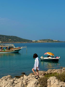 A young girl in a white dress stands on rocky terrain by a calm blue sea, with traditional boats anchored nearby.