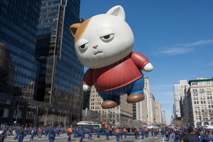 A giant inflatable cat mascot wearing a red sweater floats down a city street, held by many handlers in blue uniforms during what appears to be a parade.