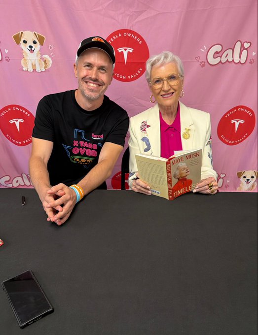 A man wearing a black cap and t-shirt sits next to an older woman with white hair and glasses who is holding her book titled 'Mave Musk' in front of a pink 'Tesla Owners Silicon Valley' backdrop featuring dog illustrations.