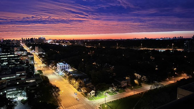 An aerial cityscape view at sunset shows streets lit by lights, with a vibrant purple and orange sky streaked with clouds.