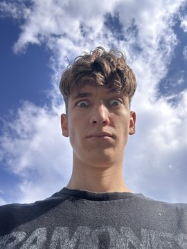 A close-up selfie shows a young man with curly brown hair making an exaggerated surprised expression, photographed from a low angle against a blue sky with scattered clouds.