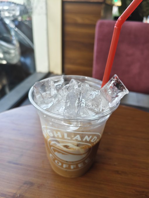 A clear plastic cup filled with iced coffee, ice cubes, and a red straw, bearing the "Highlander Coffee" logo, sitting on a wooden table.