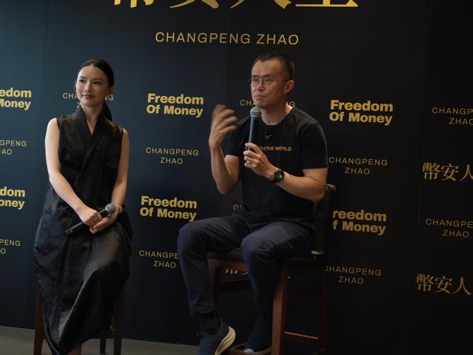 A woman and a man wearing a black "POSITIVE WORLD" t-shirt sit on stools in front of a backdrop reading "Freedom of Money" and "CHANGPENG ZHAO" (CZ), as the man speaks into a microphone. The scene corresponds to the post's playful reference to "@sisibinance" sitting next to "Big Cousin" CZ, presumably Yi He, Binance's co-founder and CZ's close associate.