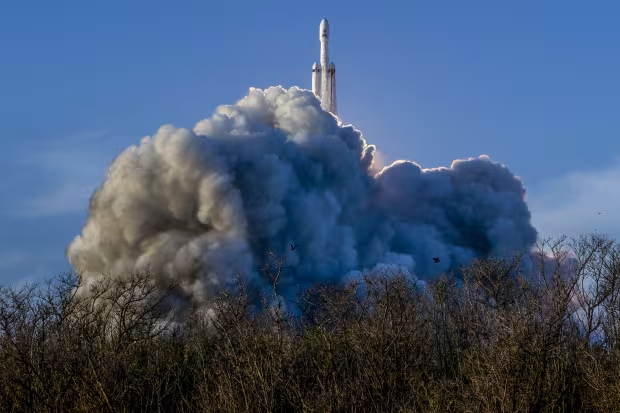 A photograph captures a rocket lifting off into a bright blue sky, with massive plumes of white and gray exhaust smoke billowing out from the launch pad behind silhouetted foliage.