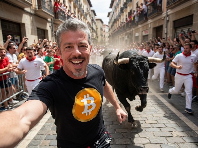 A digitally edited image of a smiling man in a black t-shirt with a Bitcoin logo running alongside a black bull through a cobblestone street lined with crowds of people, resembling the Running of the Bulls.