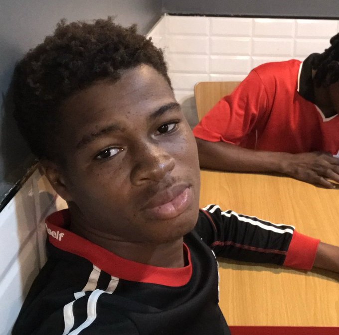 A close-up selfie of a young man with curly hair wearing a black and red jersey, with another person in a red shirt visible in the background.