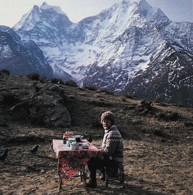 A surreal photograph of an elderly man in a patterned sweater sitting at a small table with a floral tablecloth in a mountainous, rocky landscape with snow-capped Himalayan peaks in the background.